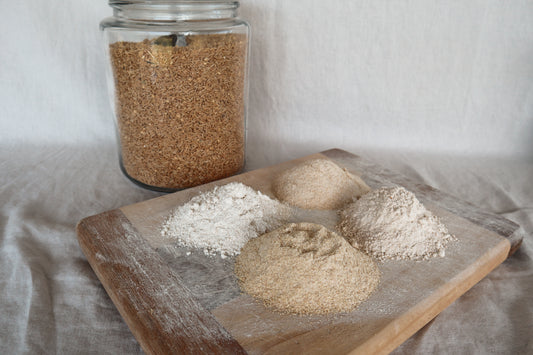 Wooden board with different types of freshly milled flour and a jar of wheat grain on a light background