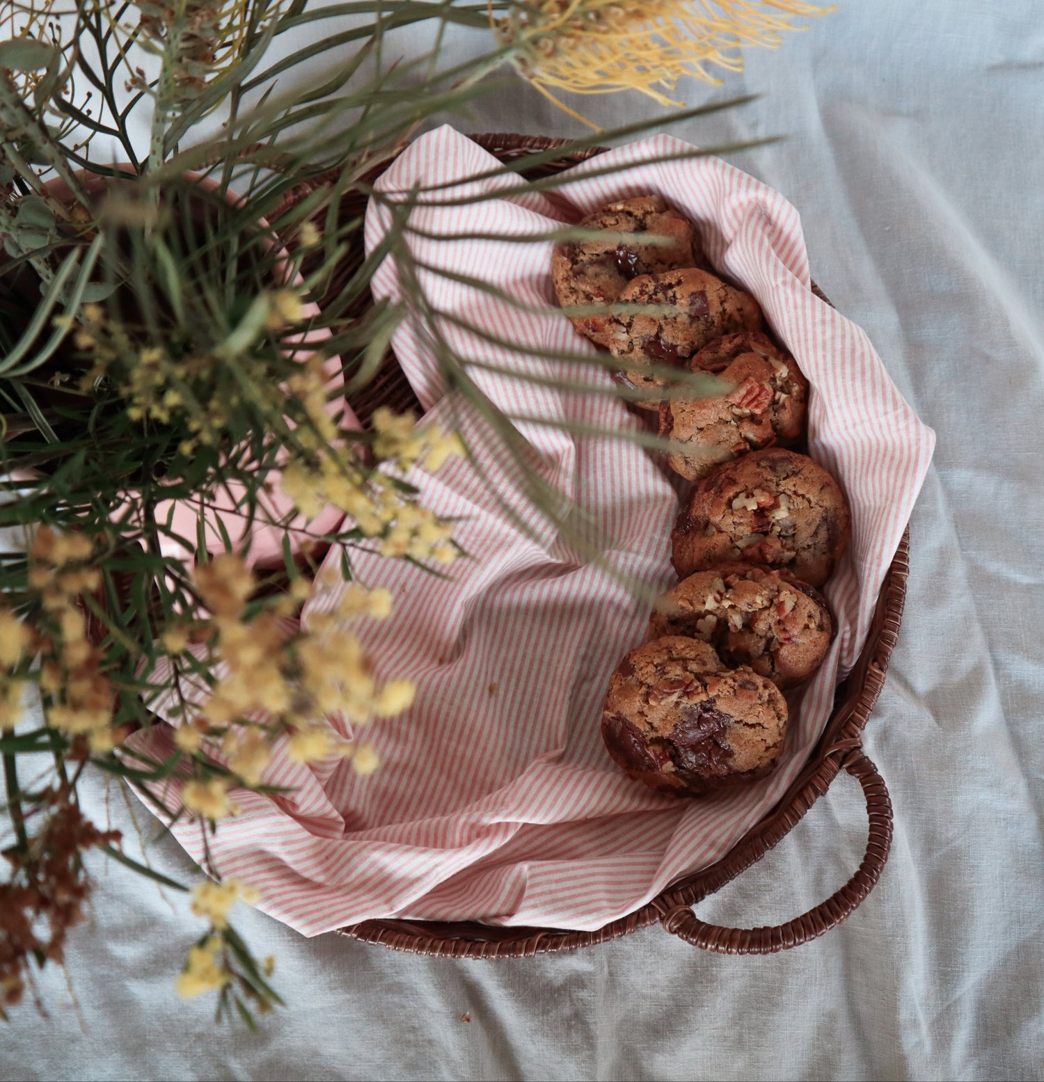 Basket of sourdough chocolate pecan cookies on a white surface with a pink jar and flowers.