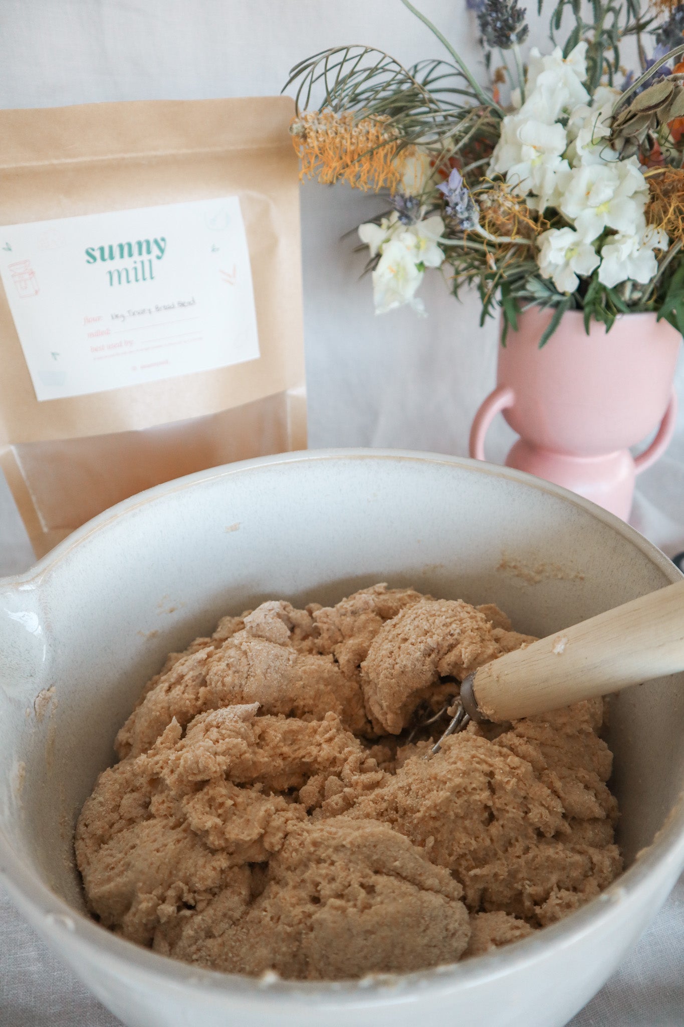 Bowl of sourdough dough with a wooden spoon, bag of flour, and flowers in the background