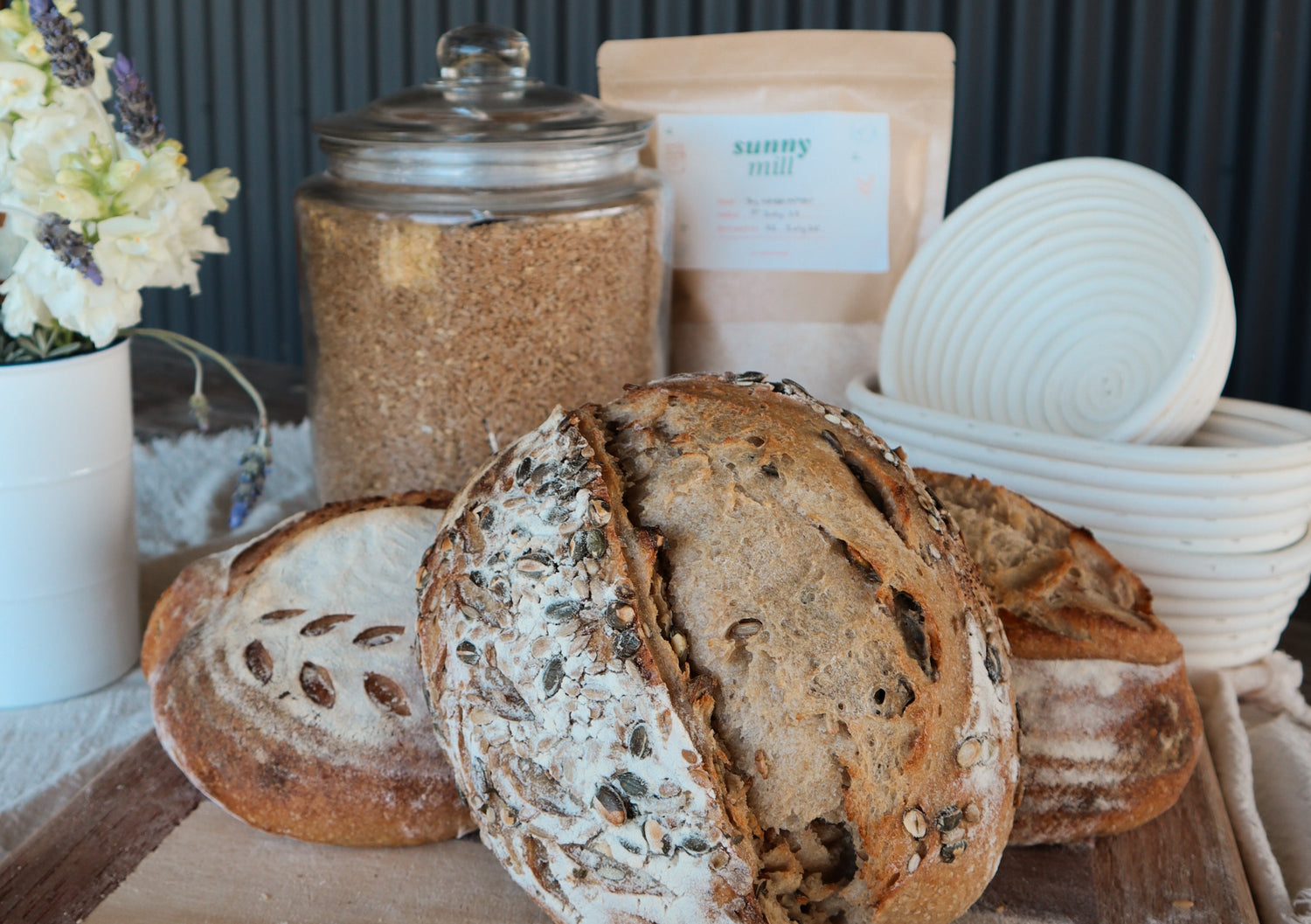 Loaves of sourdough bread on a wooden board with a jar of wheat grain and bag of freshly milled flour in the background.