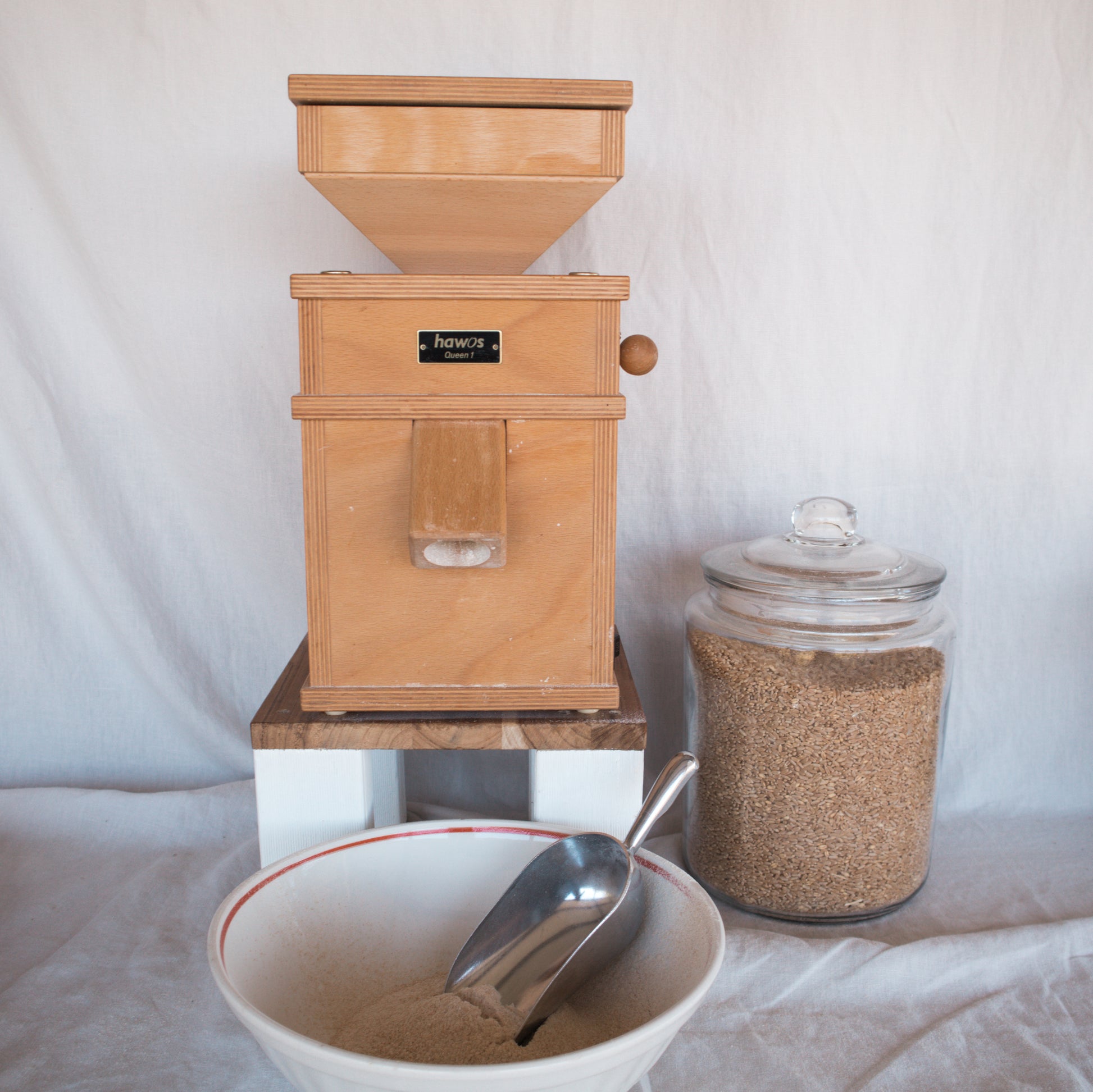 Wooden grain mill with a glass jar of wheat grain and a bowl of freshly milled wheat flour on a white background