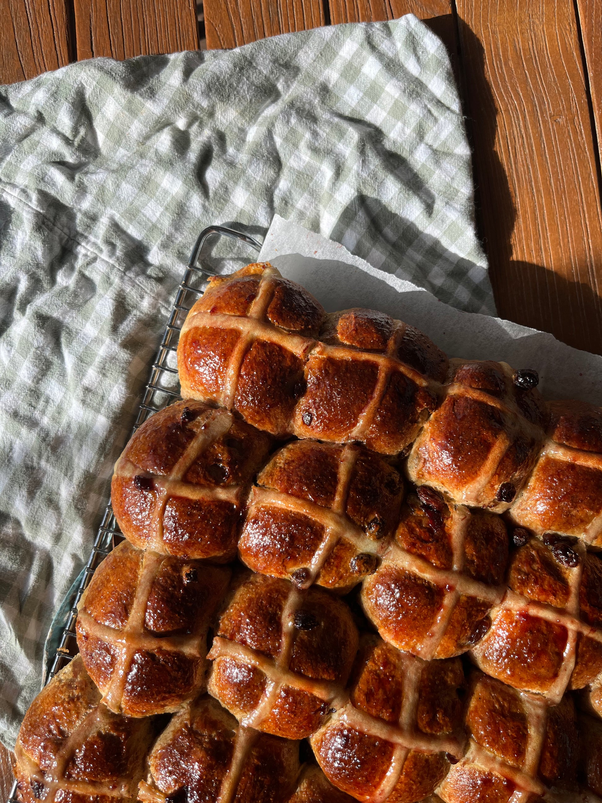 Baked hot cross buns on a cooling rack with a checkered cloth underneath.